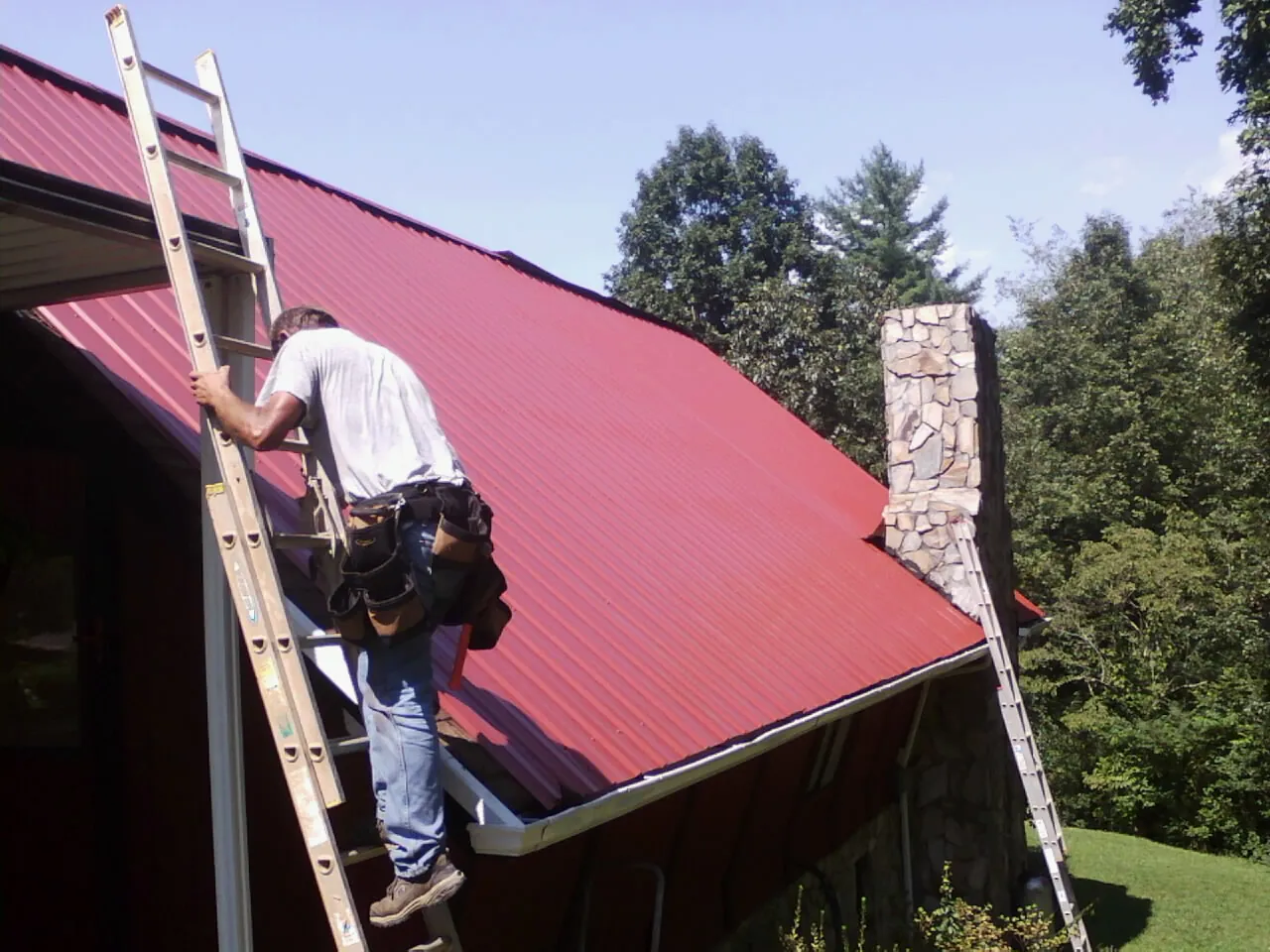 Roofer on a ladder beside a red metal roof and stone chimney — Soffit & Fascia Repair by Roofing Brothers in North Falmouth