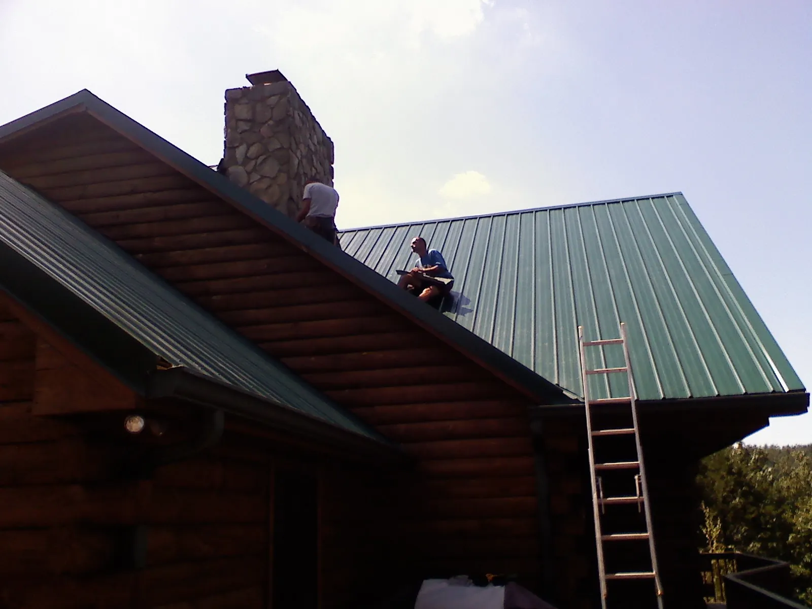 Roofers installing green metal panels beside a stone chimney on a log home in North Falmouth